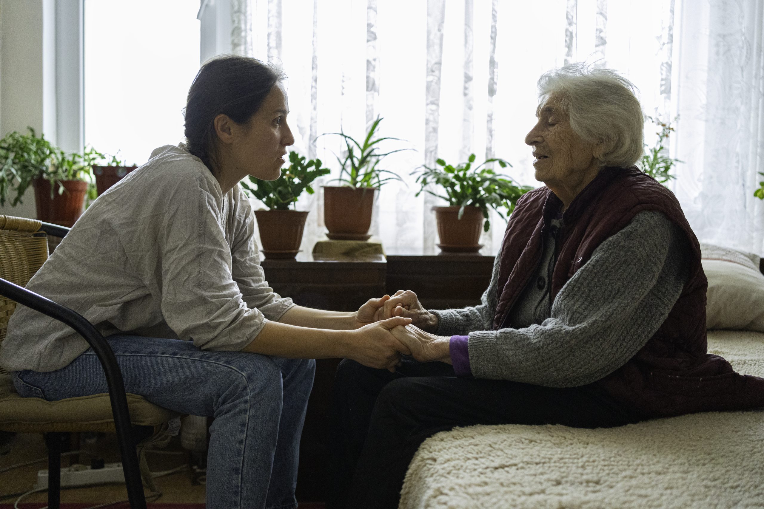 A woman holds an older woman’s hands while sitting with her and offering comfort at home.
