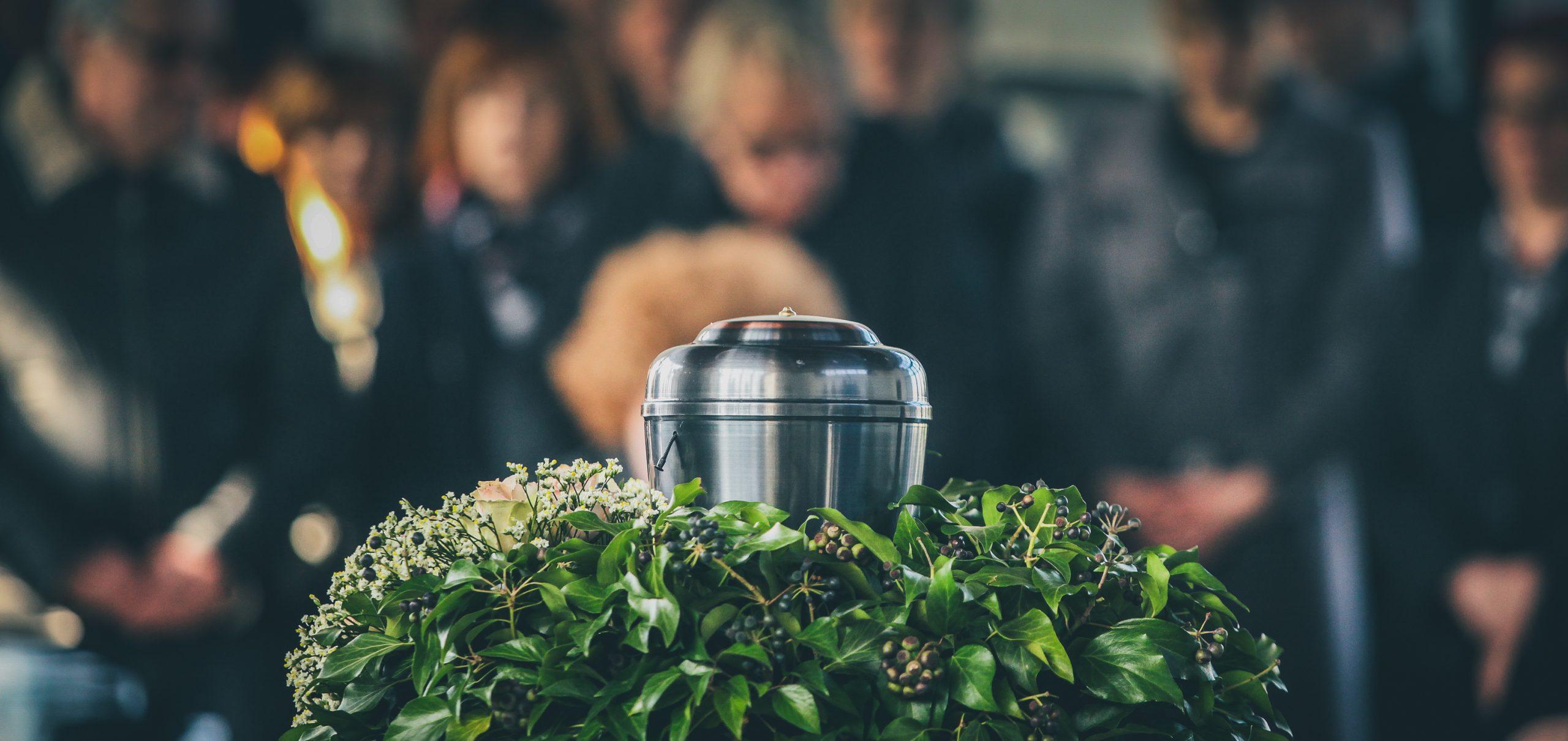A silver cremation urn surrounded by greenery and white flowers, with mourners gathered in the softly blurred background.